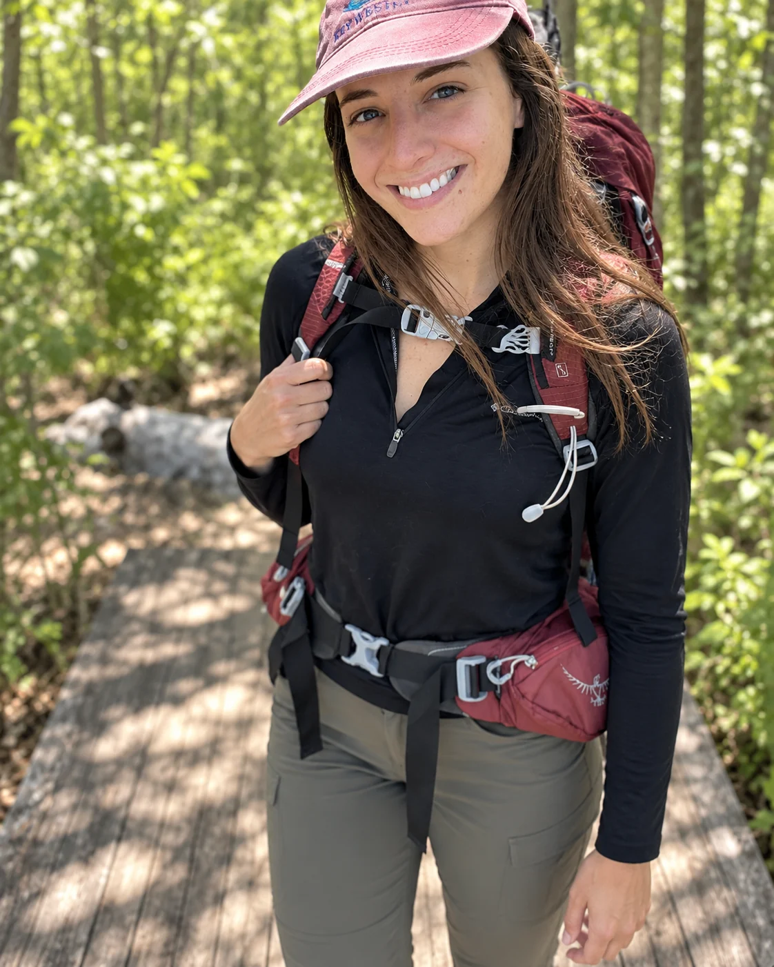 Abbey smiling in a forest, wearing a pink cap and backpack